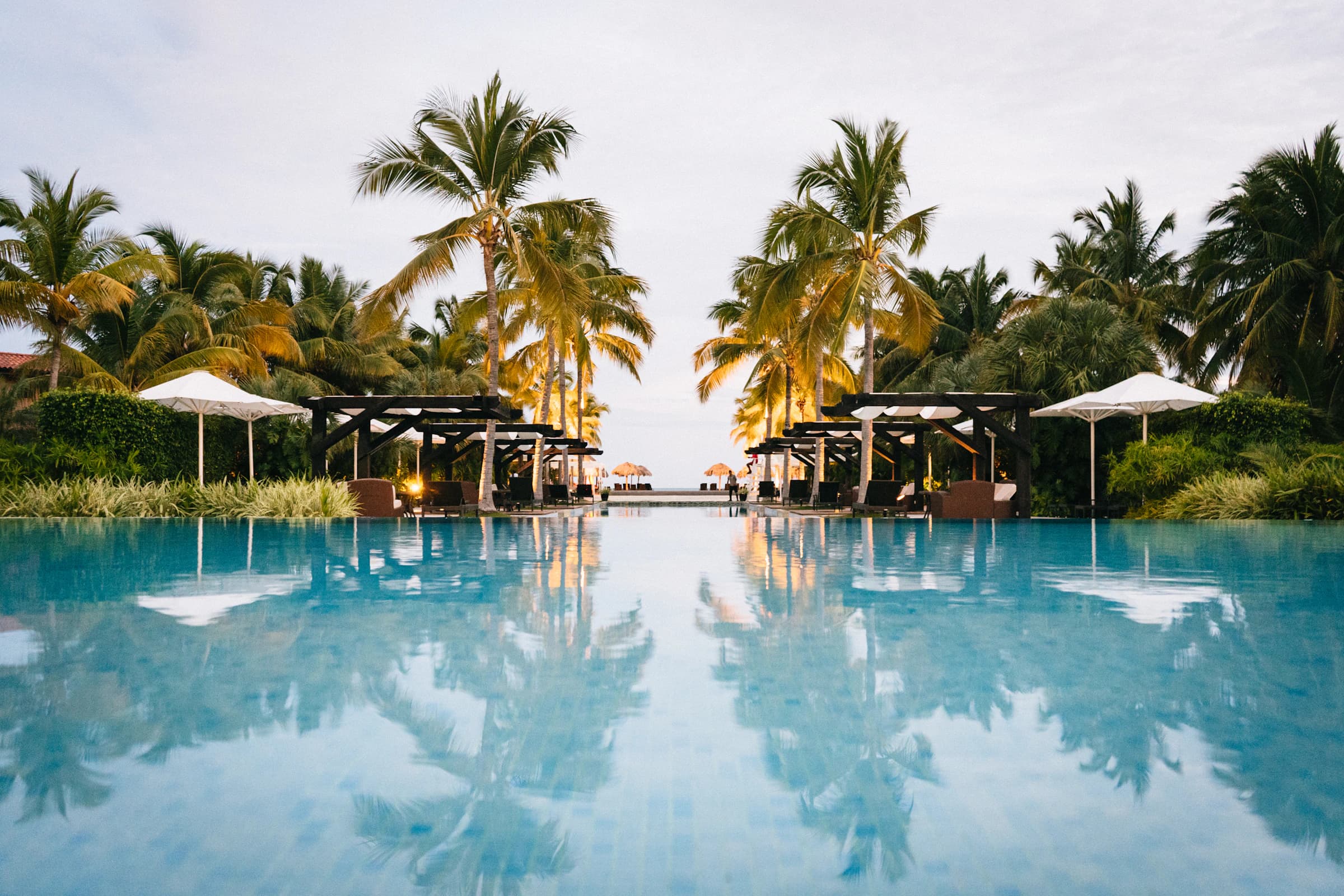 Swimming pool with endless edge mirroring palm trees and white umbrellas in golden hour, with the sea at the bottom